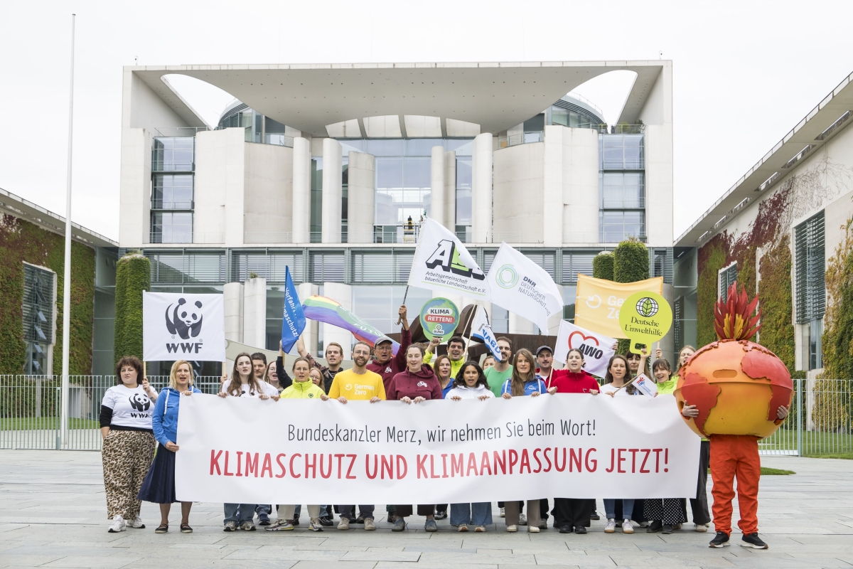 zwei Dutzend Umweltaktive vor dem Bundeskanzleramt in Berlin mit einem Banner, Aufschrift "Bundeskanzler Merz, wir nehmen Sie beim Wort! KLIMASCHUTZ UND KLIMAANPASSUNG JETZT!"; rechts im Bild, eine als brennende Erde verkleidete Person